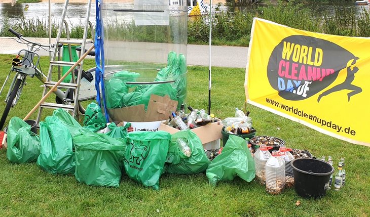 Zahlreiche gefüllte Müllbeutel liegen für ein Foto aufgestapelt vor einer Fahne mit der Aufschrift World Cleanup Day. Außerdem sind mehrere Flaschen zu sehen, die mit gesammelten Zigarettenkippen voll sind.