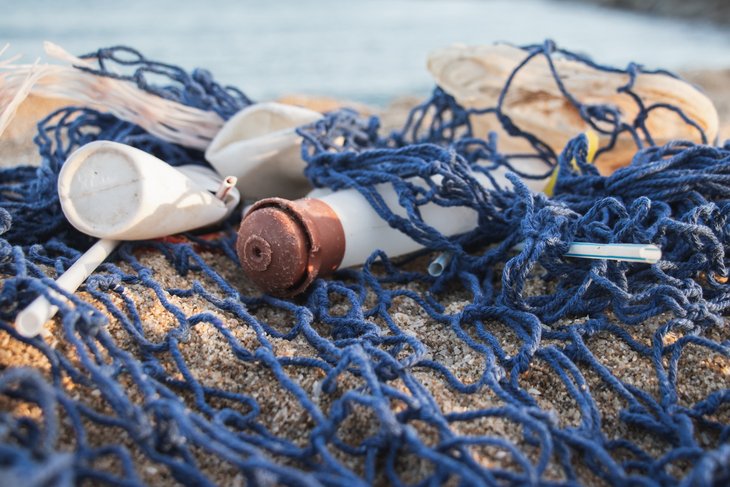 Eine Plastikflasche, ein Strohhalm und weitere Plastikreile hängen in einem Fischernetz, das am Strand liegt.